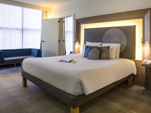 A hotel room with a large bed, a bedside table with a phone, a wall-mounted light, and a seating area with a couch and curtains. - Novotel Geneve Centre (Geneva)