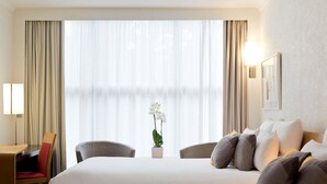 A hotel room with two beds, a desk, a chair, a lamp, and a window with curtains. - Novotel Geneve Centre (Geneva)