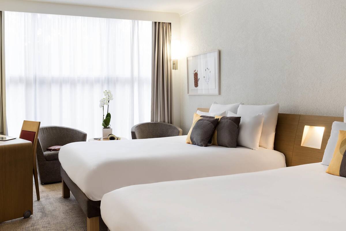 Habitación de hotel con dos camas, un escritorio, sillas, una planta en maceta y un cuadro en la pared.