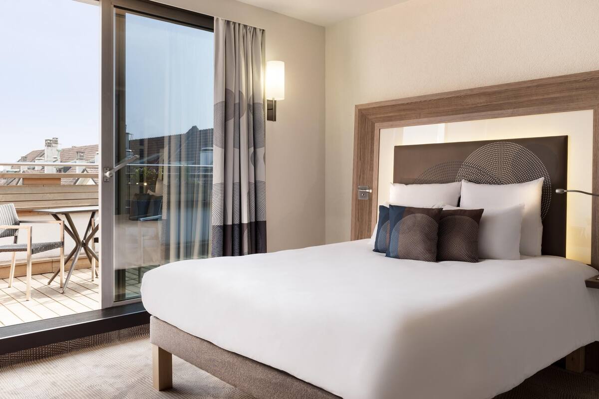 Una habitación de hotel con una cama grande, un puerta de vidrio que da a un balcón y un cabecero moderno.