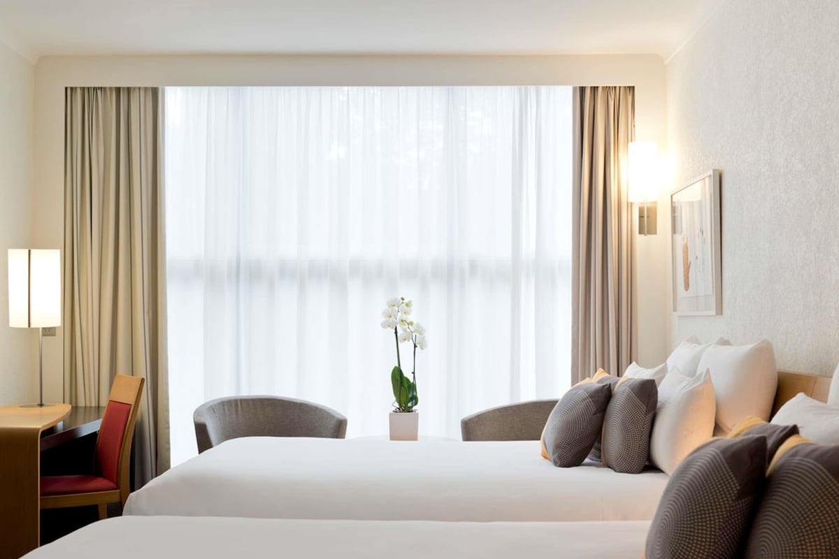 Une chambre d’hôtel avec deux lits, un bureau, une chaise, une lampe et une fenêtre avec des rideaux.