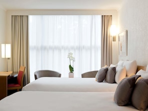 Habitación de hotel con dos camas, un escritorio, una silla, una lámpara y un ventanal con cortinas.