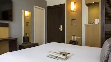 A hotel room with a bed, a flat-screen TV mounted on the wall, a wooden door, a wall-mounted light, and a small shelf with a cup and a mug.