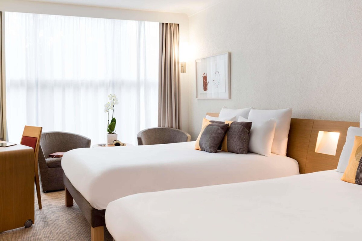 Ett hotellrum med två sängar, ett skrivbord, stolar, en krukväxt och ett fönster med gardiner.