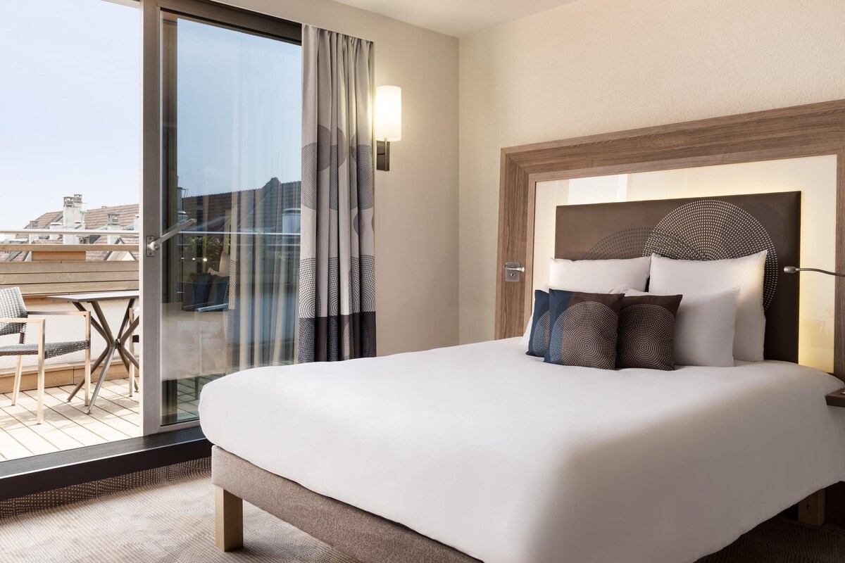 Une chambre d’hôtel avec un grand lit, une porte vitrée donnant sur un balcon et une tête de lit moderne.