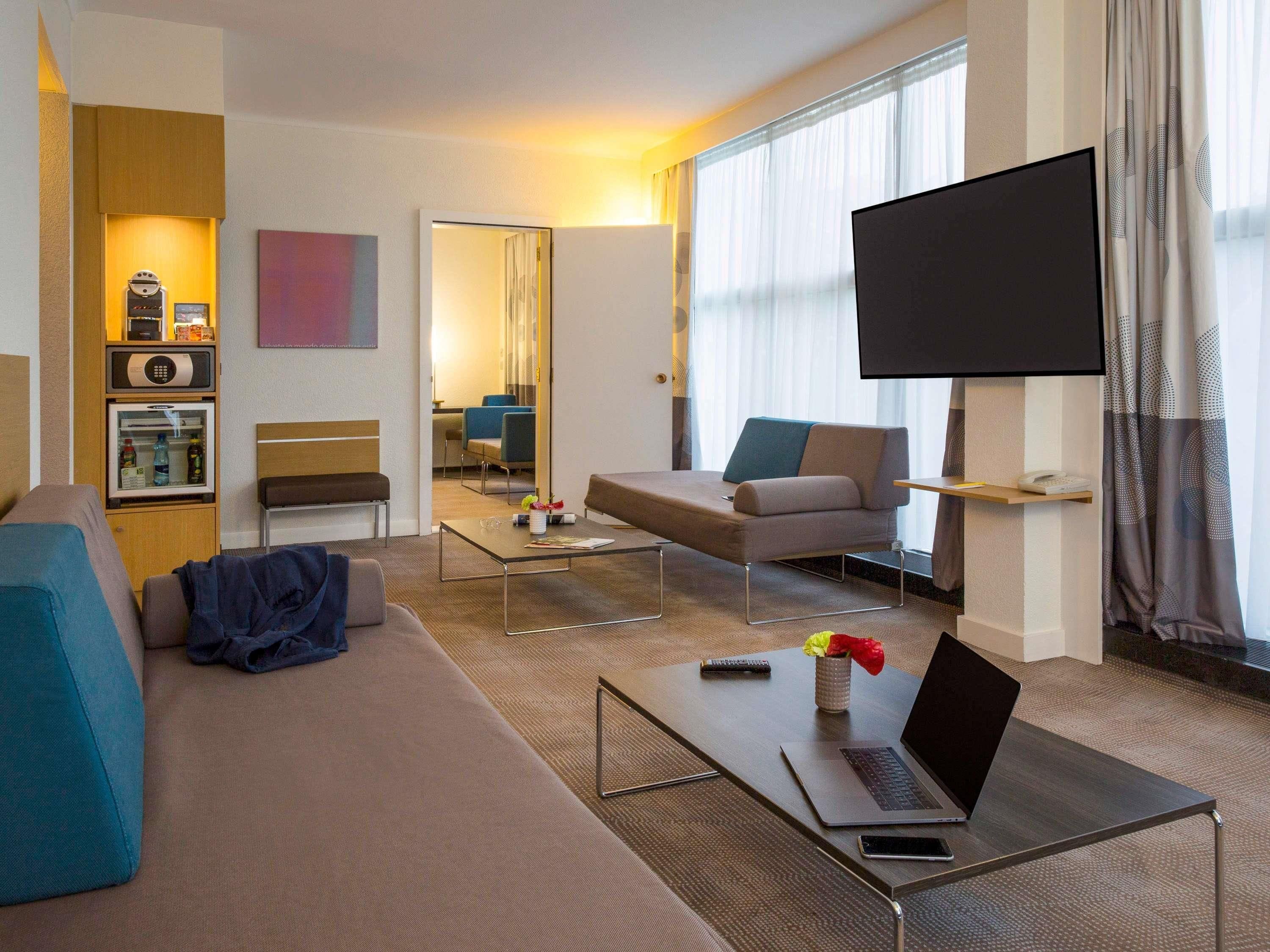 A modern hotel room with a sofa, a coffee table, a TV, and a minibar.