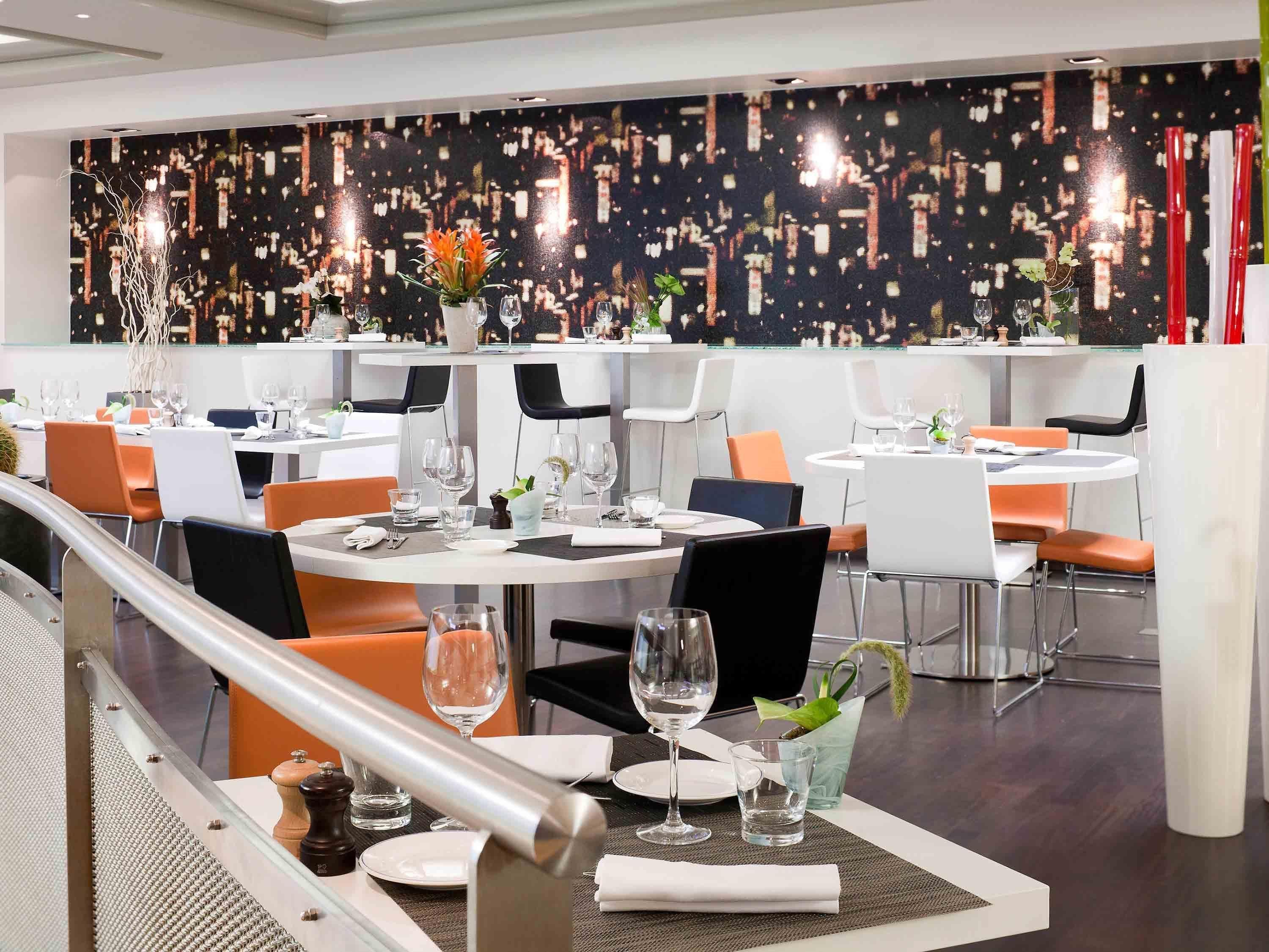 Un restaurant moderne avec des tables rondes, des chaises orange et noires, et une fresque représentant un paysage urbain sur le mur.