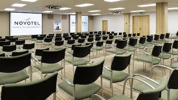 A conference room with rows of chairs, a projector screen displaying 'NOVOTEL HOTELS, SUITES & RESORTS', and a large door.