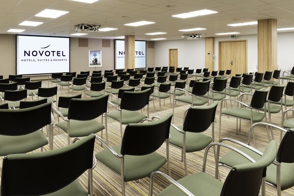 A conference room with rows of chairs, a projector screen displaying 'NOVOTEL HOTELS, SUITES & RESORTS', and a large door.