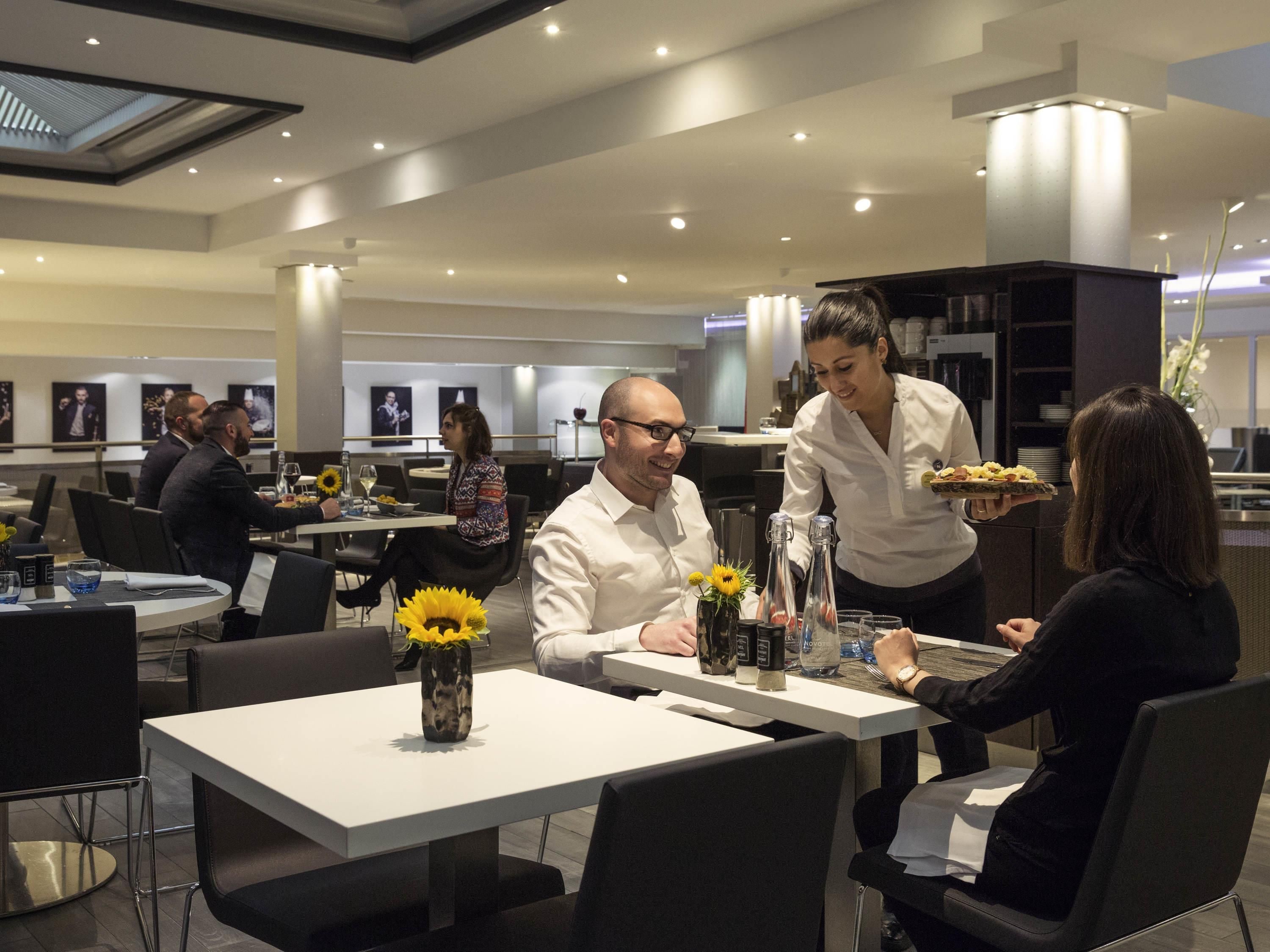 A modern dining area with white tables and black chairs, featuring a central vase with yellow flowers.