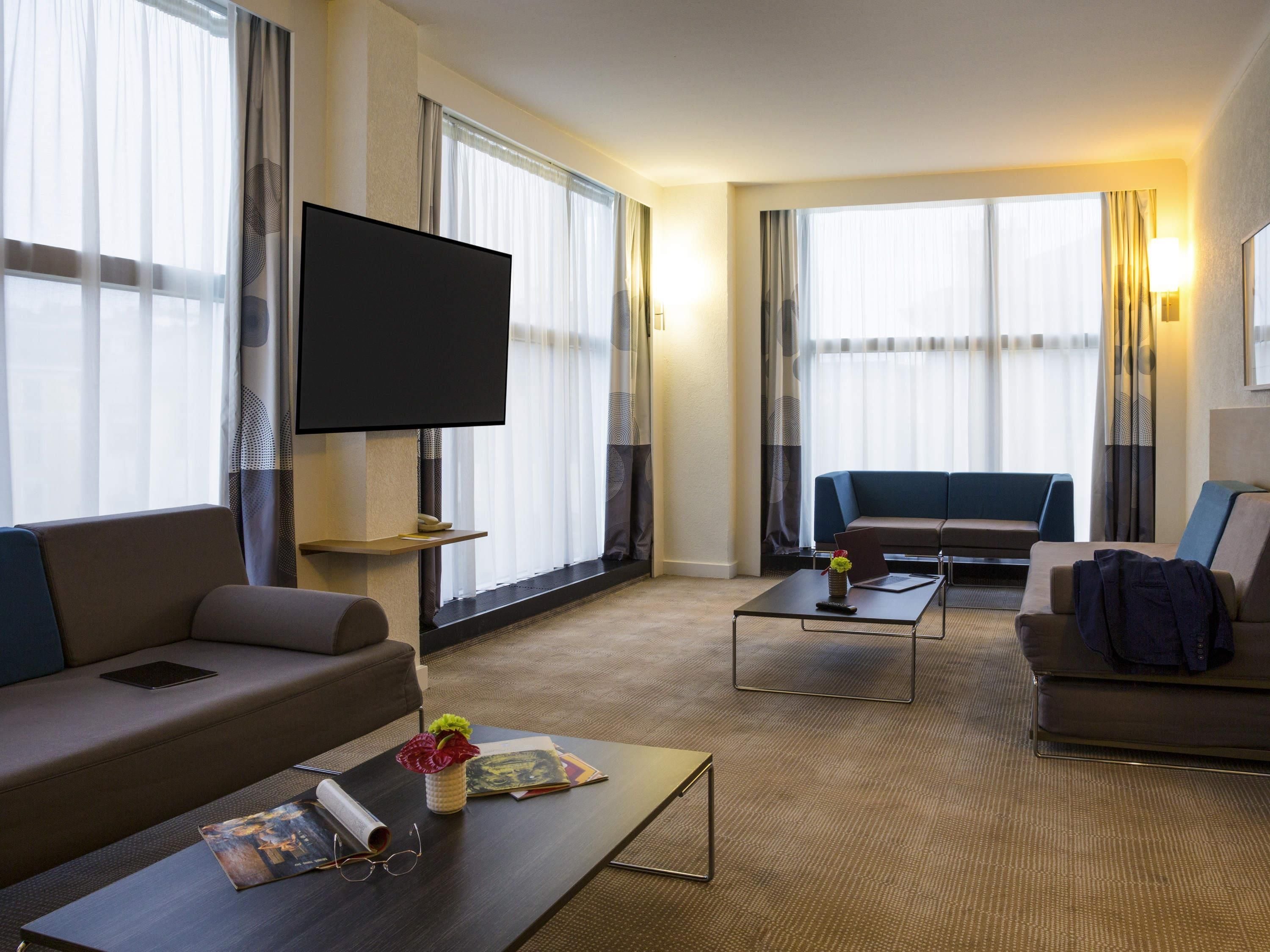 Habitación de hotel con televisión de pantalla plana, dos sofás, una mesita de centro y ventanas amplias con cortinas.