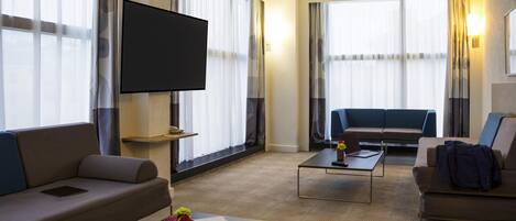 Habitación de hotel con televisión de pantalla plana, dos sofás, una mesita de centro y ventanas amplias con cortinas.