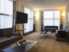 A hotel room with a flat-screen TV, two sofas, a coffee table, and large windows with curtains. - Novotel Geneve Centre (Geneva)