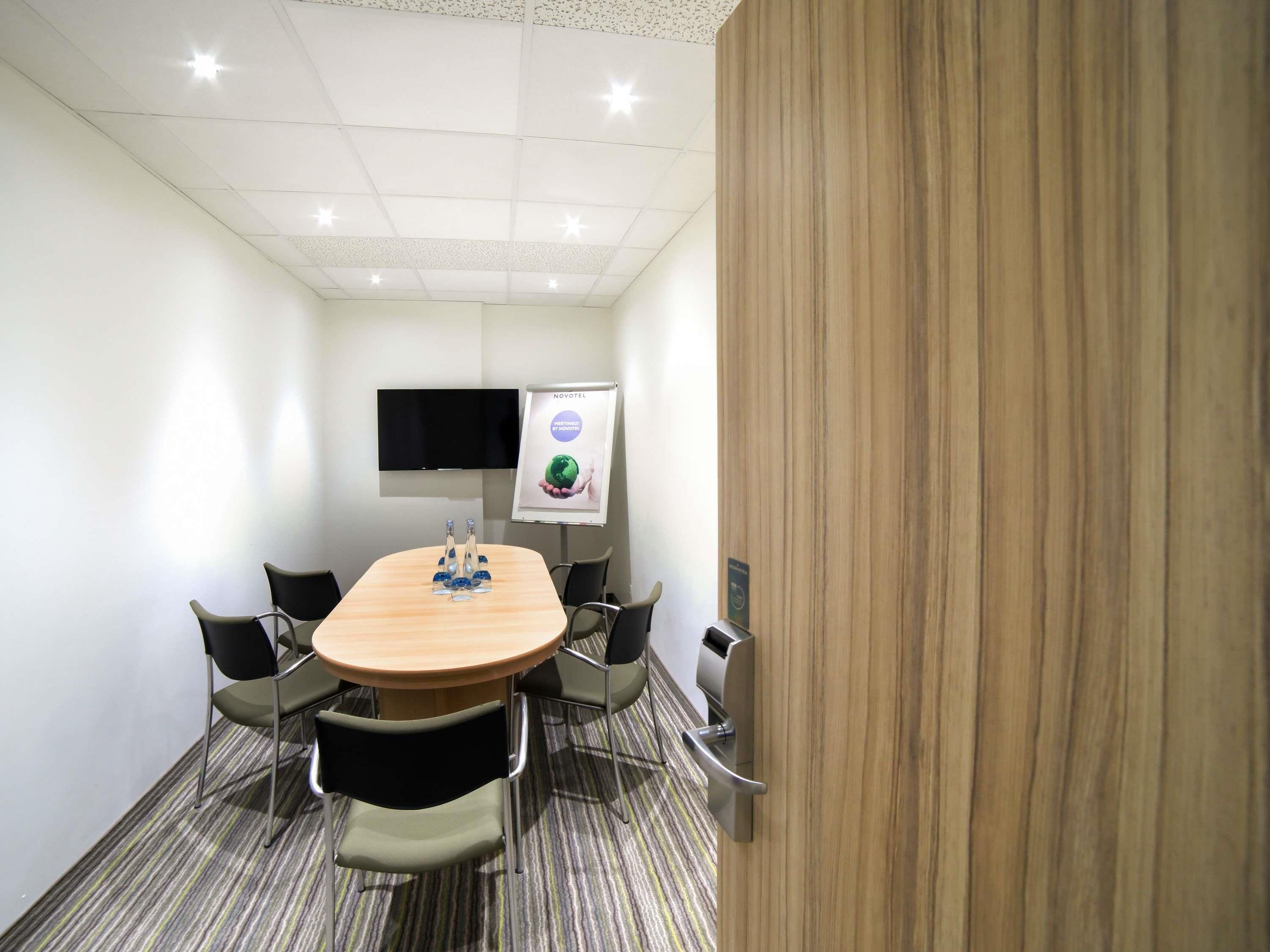 A conference room with a large oval table, six chairs, a flat-screen TV, and a poster on the wall.