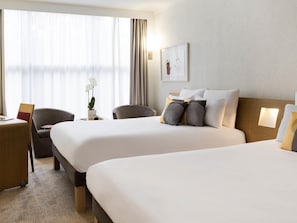 A hotel room with two beds, a desk, chairs, a potted plant, and a framed picture on the wall. - Novotel Geneve Centre (Geneva)