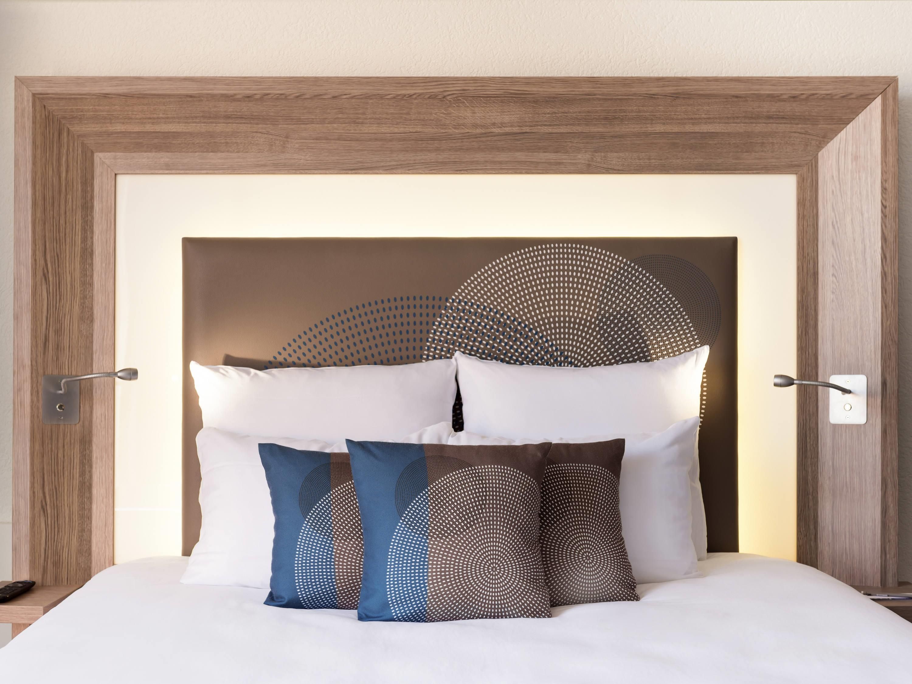 Una cama bien hecha con ropa blanca y almohadillas decorativas, cabecera de madera y lámpara de pared.