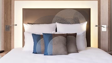 Una cama bien hecha con ropa blanca y almohadillas decorativas, cabecera de madera y lámpara de pared.