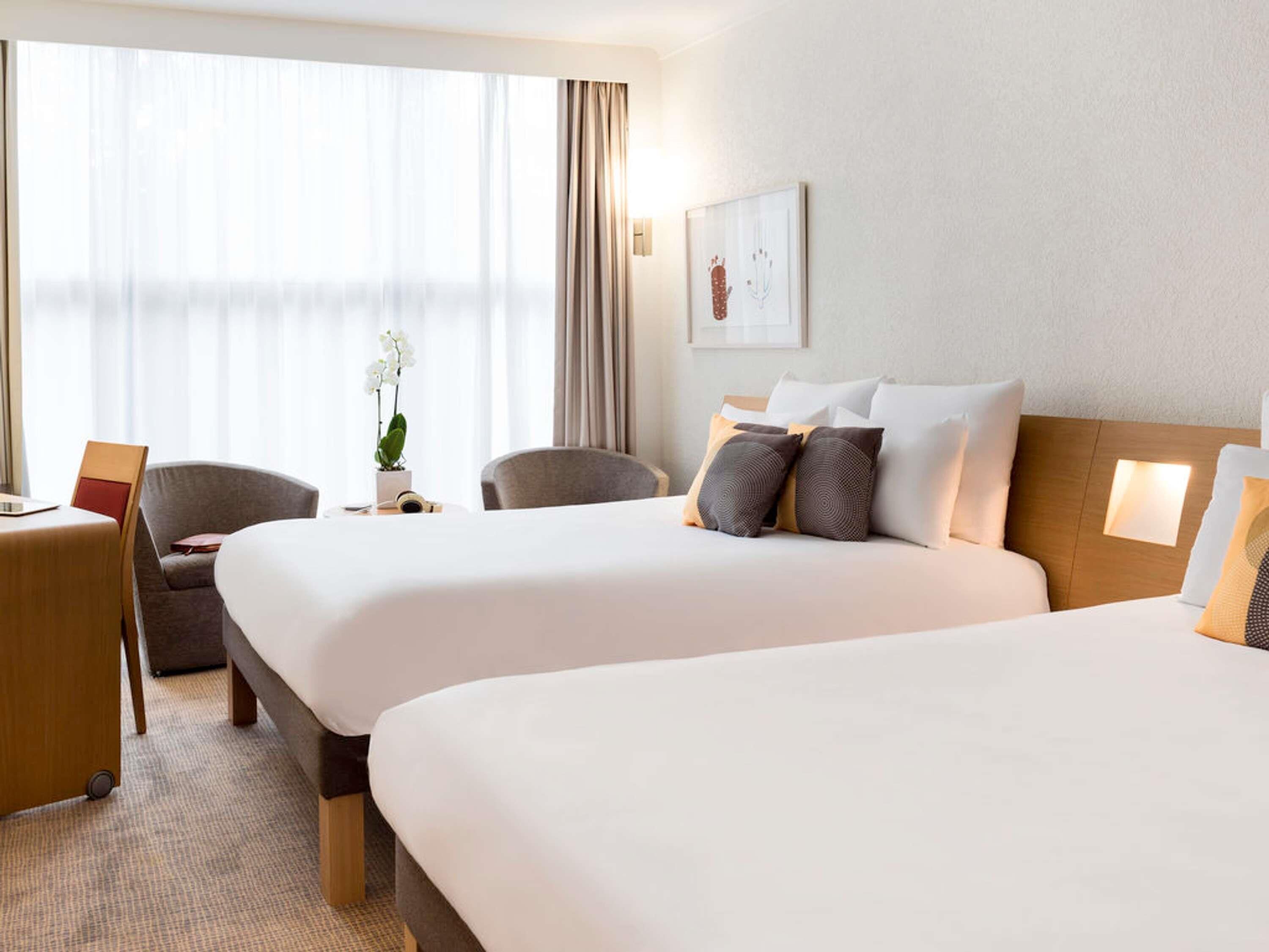 Ett hotellrum med två sängar, ett skrivbord, stolar, en krukväxt och ett fönster med gardiner.