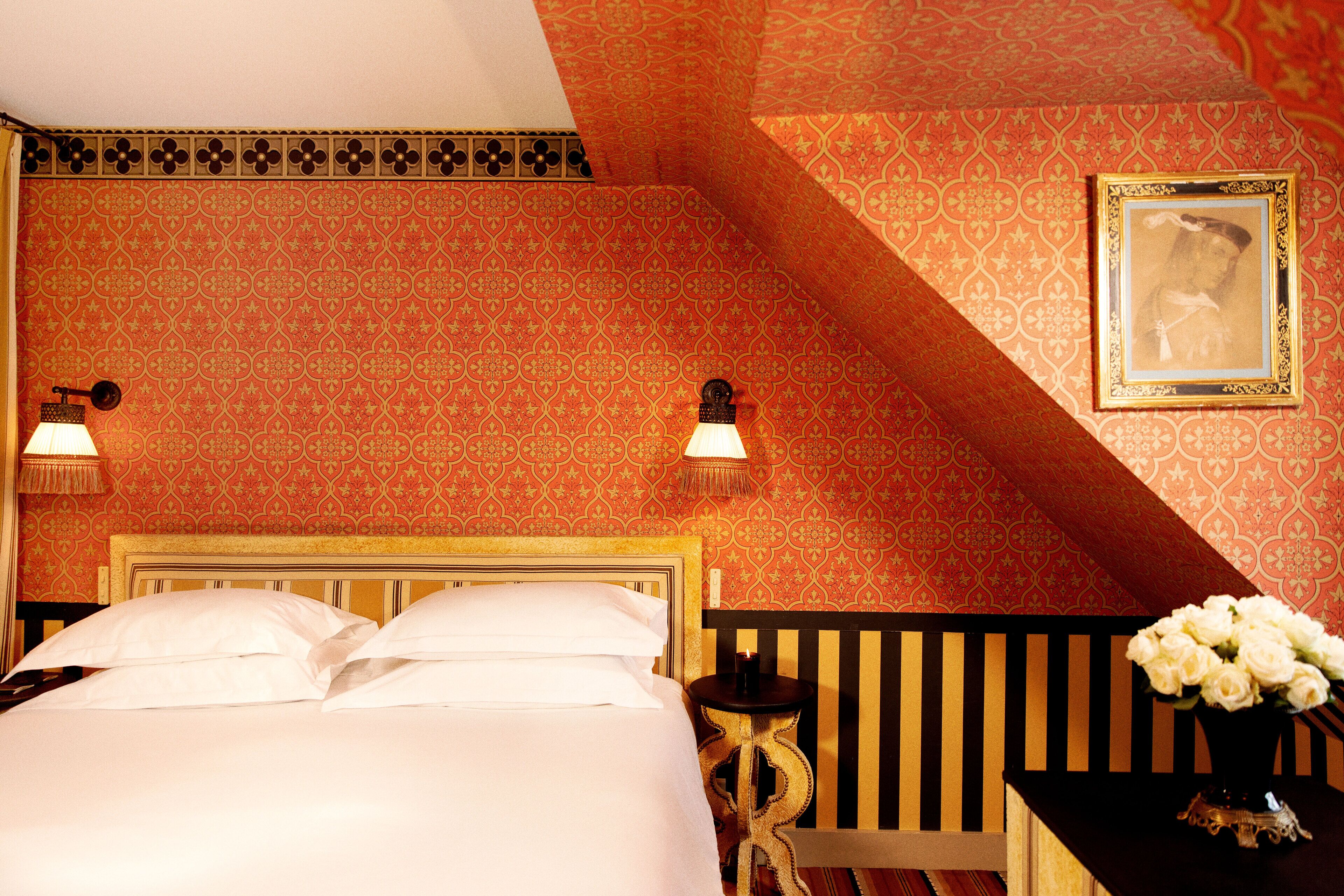 Habitación de hotel con una cama, dos lámparas de pared, una mesita de noche, una pequeña mesa con un florero y un cuadro en la pared.