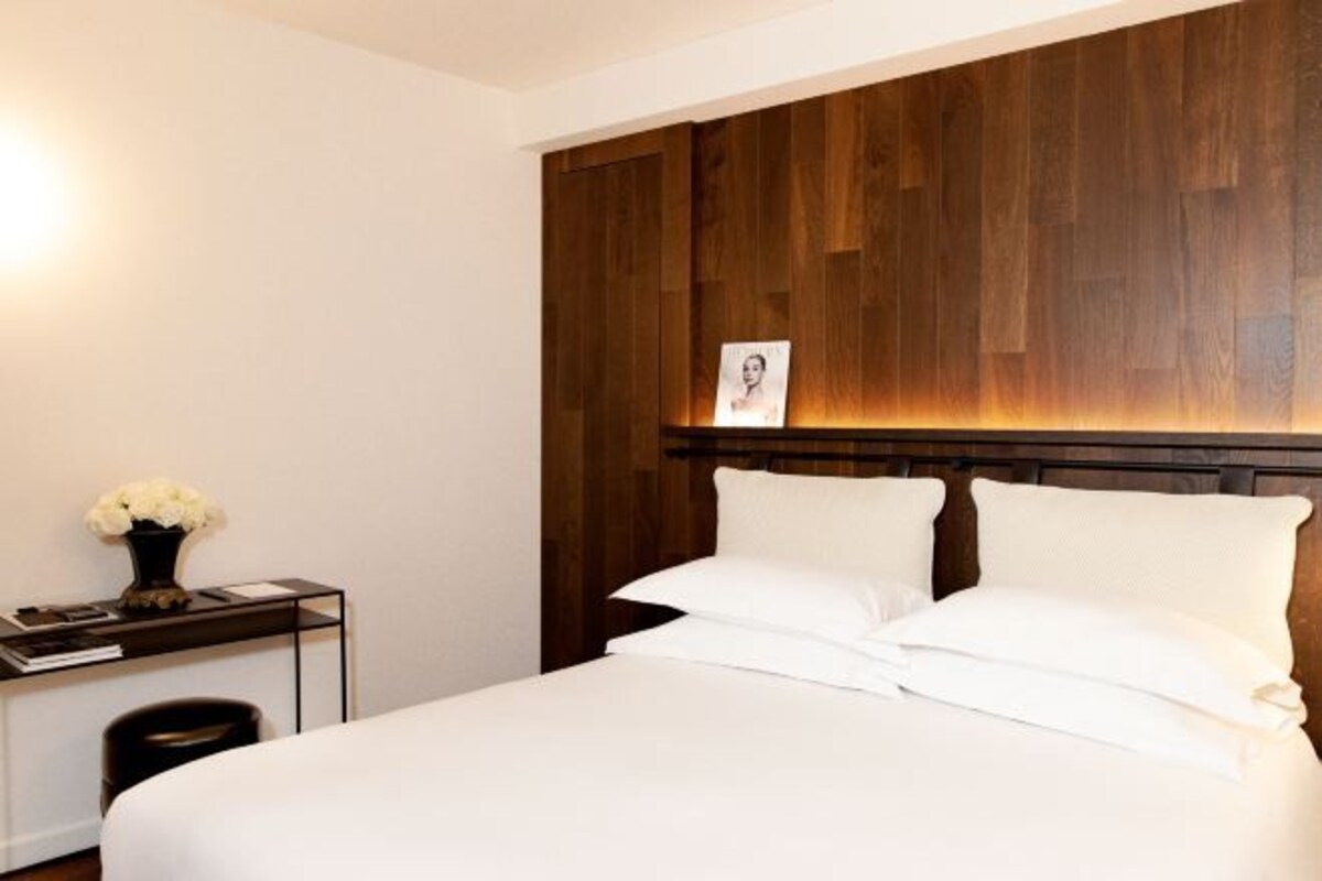 Habitación de hotel con cabecera de madera, cama con ropa blanca impecable, mesita de noche con un florero y un cuadro enmarcado en la cabecera.