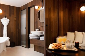A modern bathroom with a wooden wall, a sink, and a mirror. There is a towel rack with a white towel, a trash bin, and a tray with croissants and orange juice.