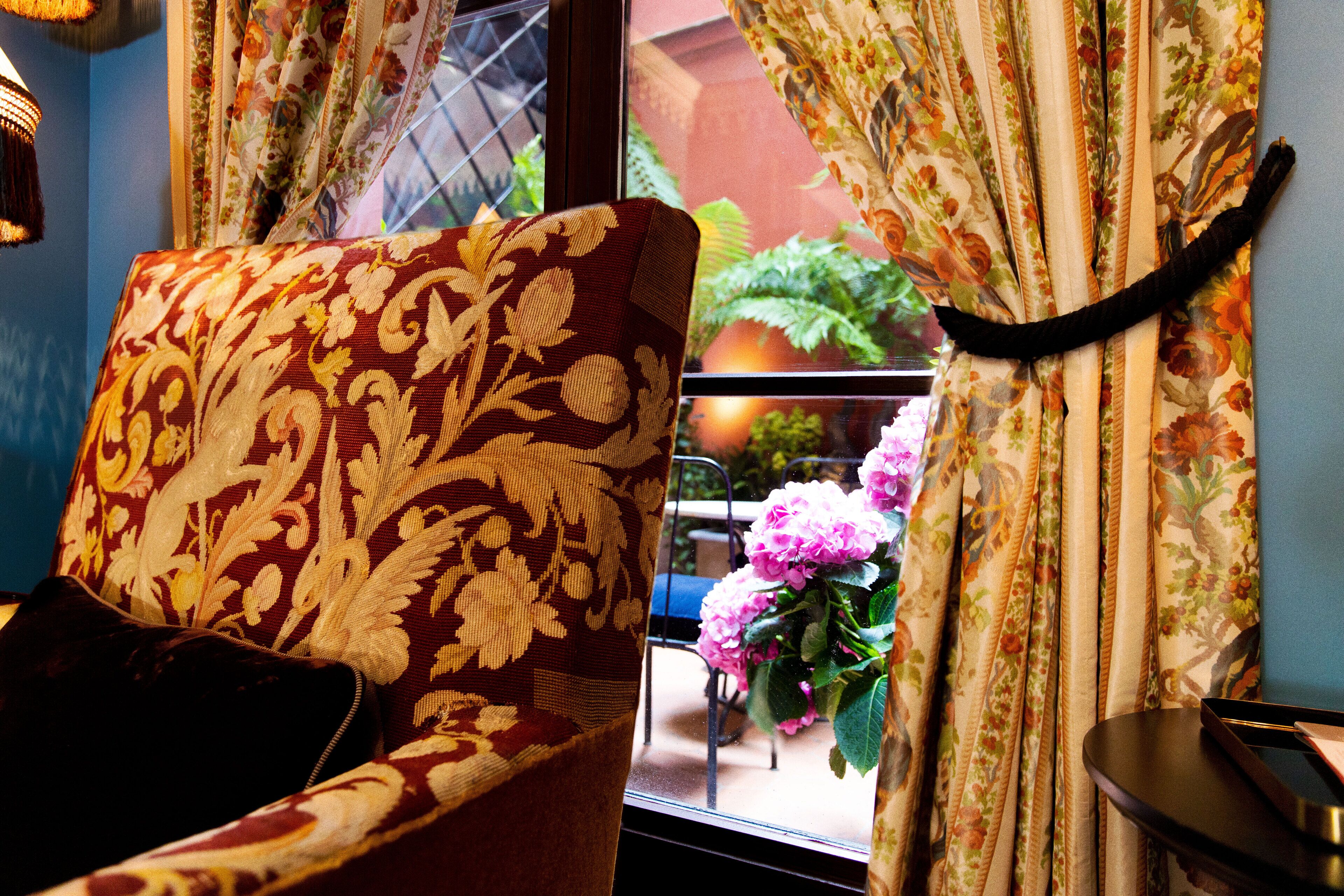 A room with a patterned armchair, a window with floral curtains, and a view of greenery outside.