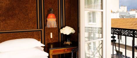 A hotel room with a bed, a nightstand with a vase of flowers, a chair, and a view of the city through a window.