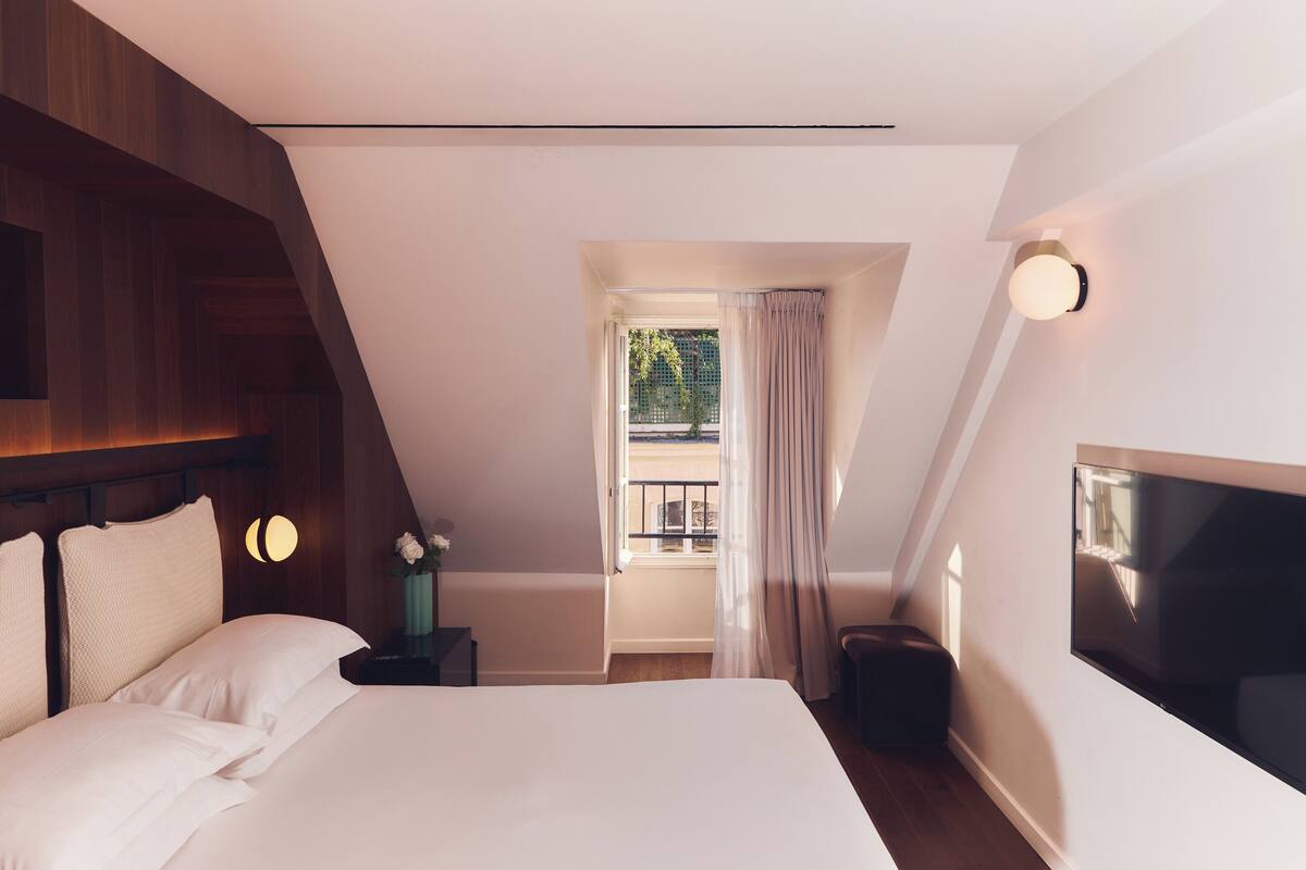 A bedroom with a large bed, a nightstand with a vase of flowers, a window with curtains, and a wall-mounted light fixture.