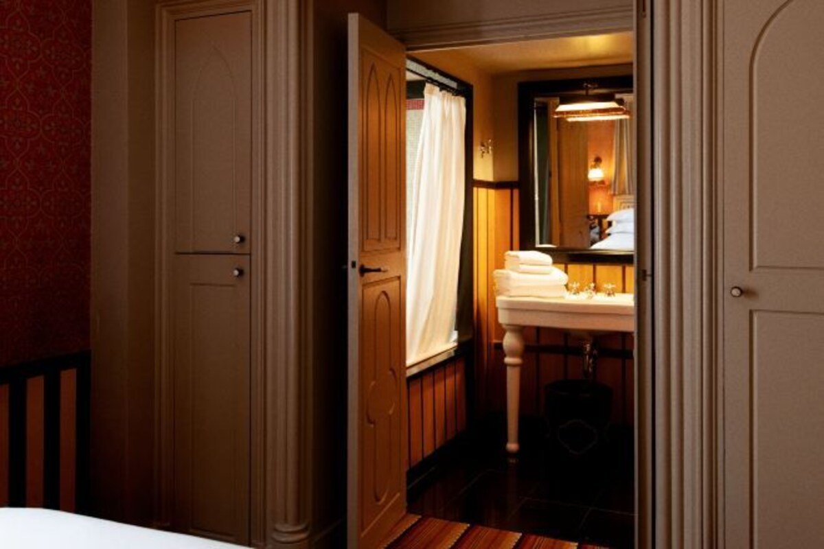 A hotel room with a wooden wardrobe, a bathroom with a sink and mirror, and a bed with white bedding.