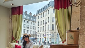 Une personne est assise sur un canapé à motifs floraux, agrémenté de coussins rouges, à côté d’une armoire en bois, dans une pièce offrant une vue sur le paysage urbain grâce à de grandes fenêtres ornées de rideaux rouges et verts.