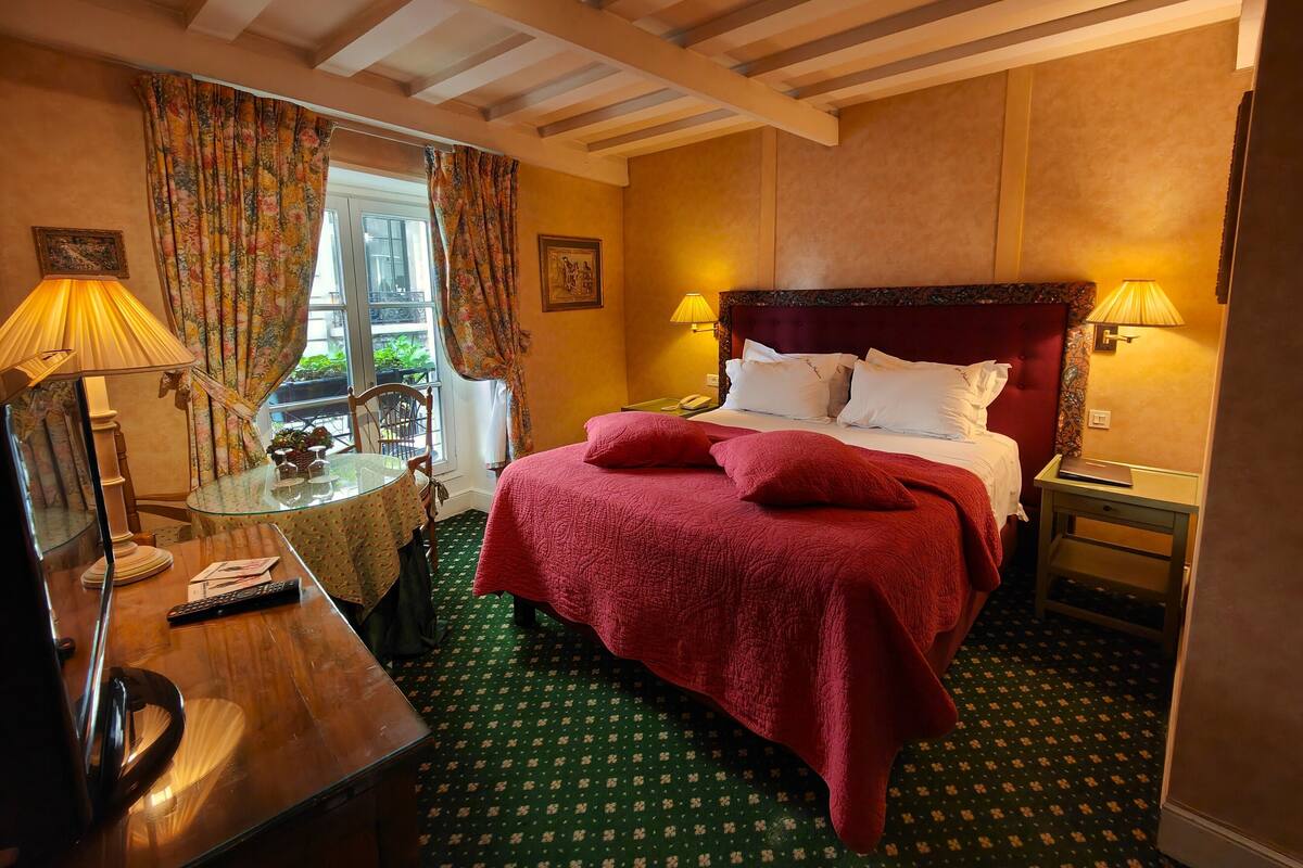 A hotel room with a bed, two bedside lamps, a glass table with a chair, a wooden desk, and a window with curtains.