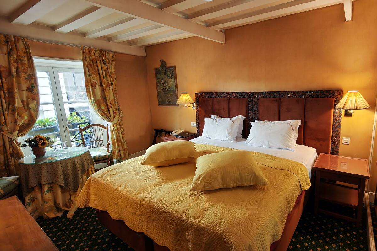 A hotel room with a large bed, two bedside lamps, a nightstand, a chair, a table with a vase of flowers, and a window with curtains.