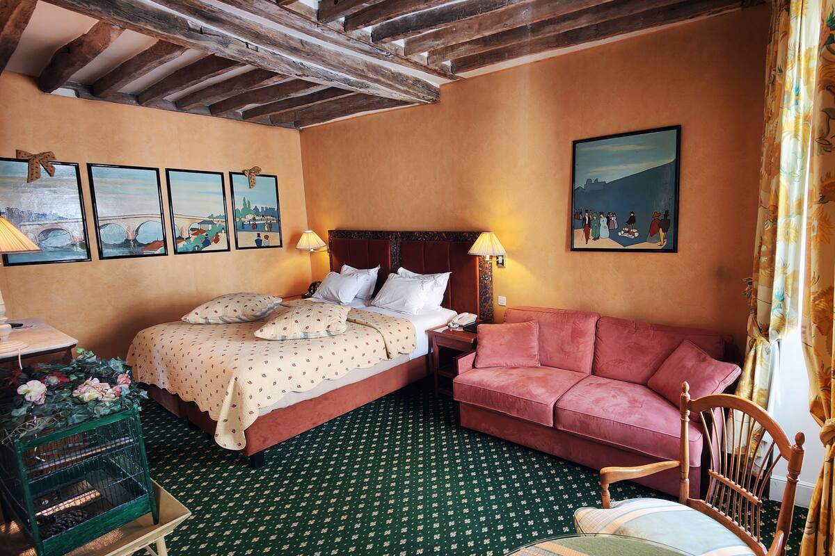 A room with two beds, a sofa, a wooden chair, a green patterned carpet, and a wall with framed artwork.