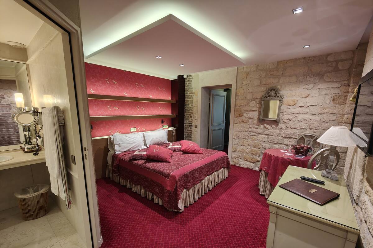 A bedroom with a red carpet, a bed with red bedding, a desk with a lamp, and a stone wall.