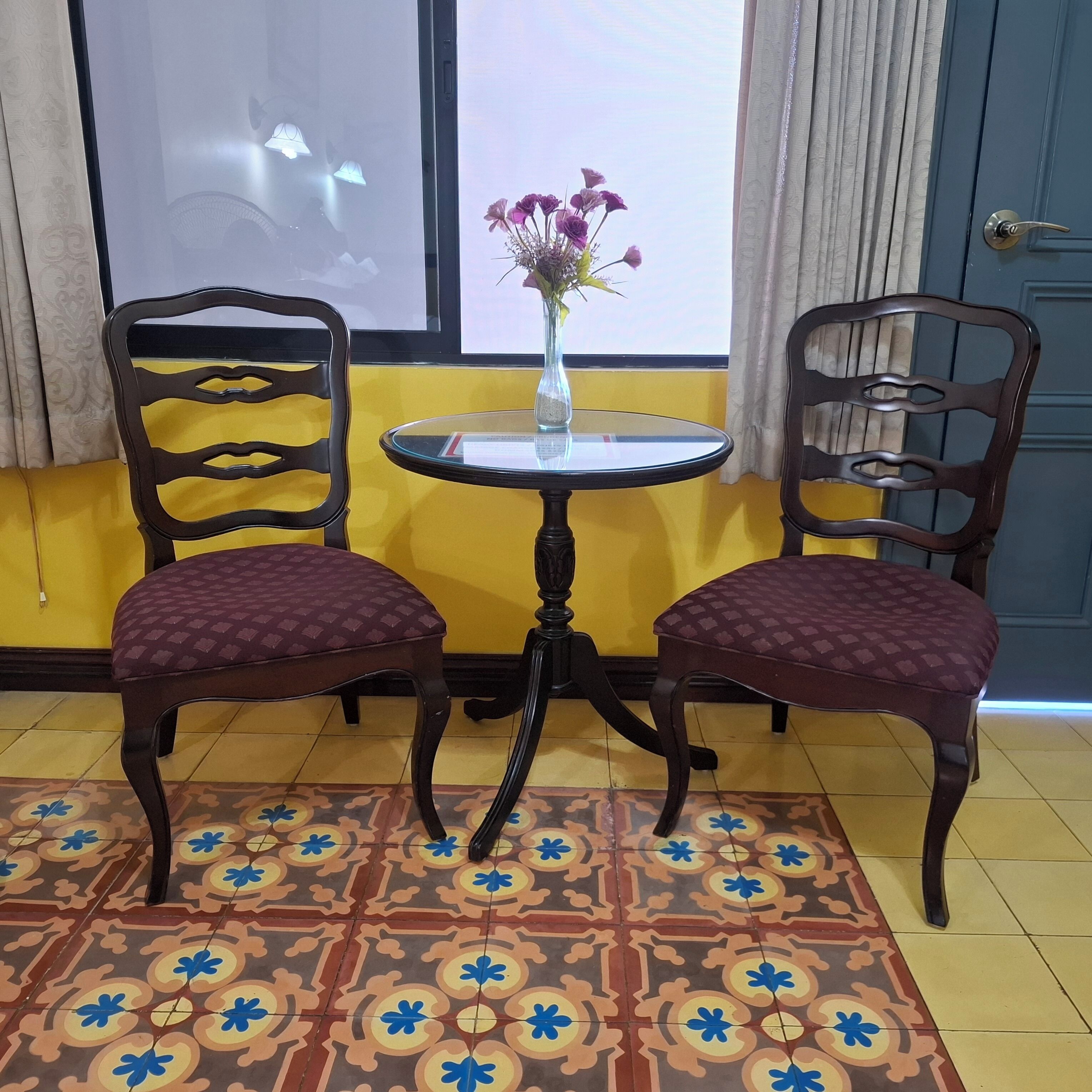 A room with two chairs, a small table, a window with curtains, and a patterned floor.