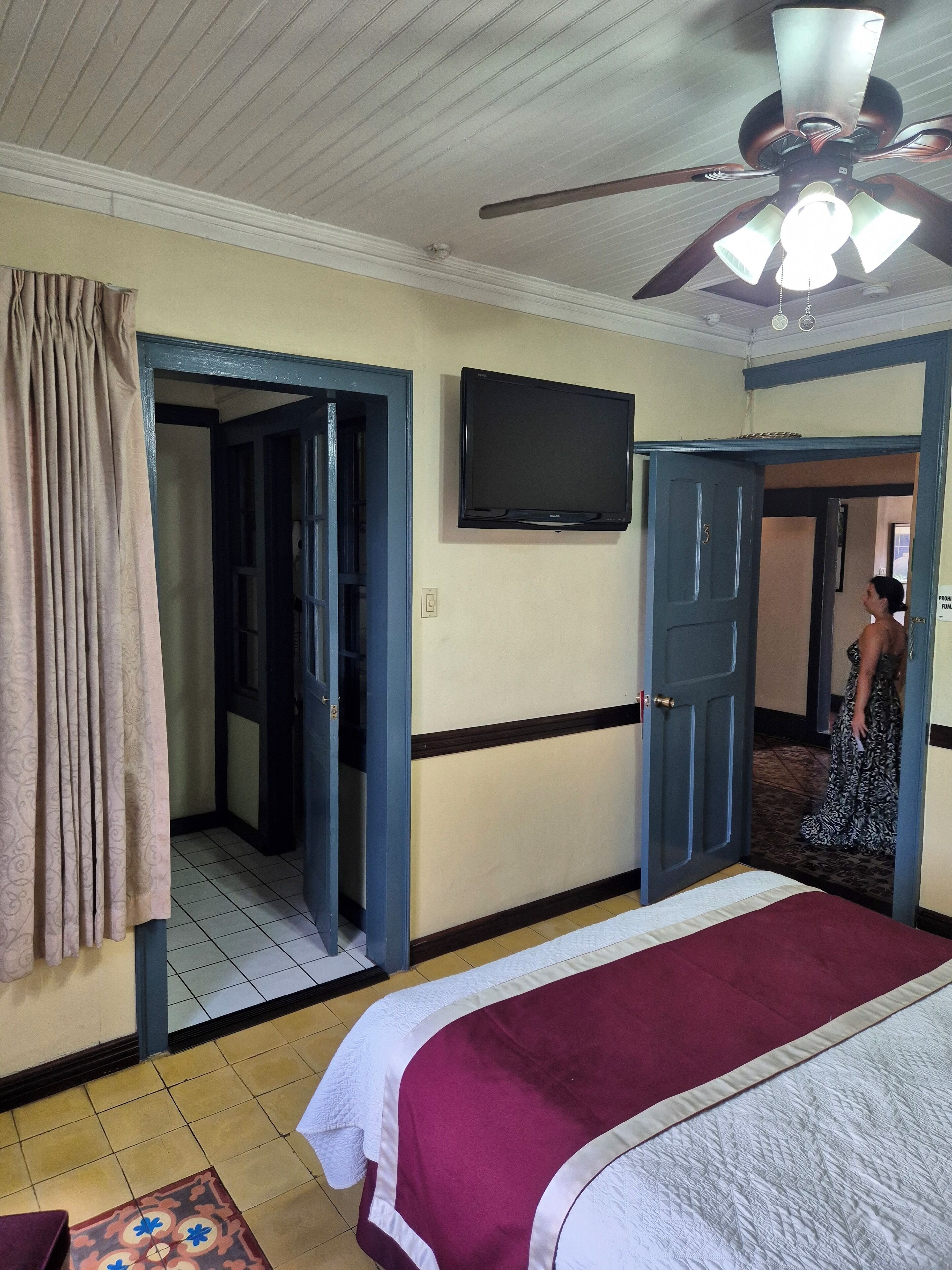 Een hotelkamer met een bed, een plafondventilator, een televisie en een deur naar een andere kamer.