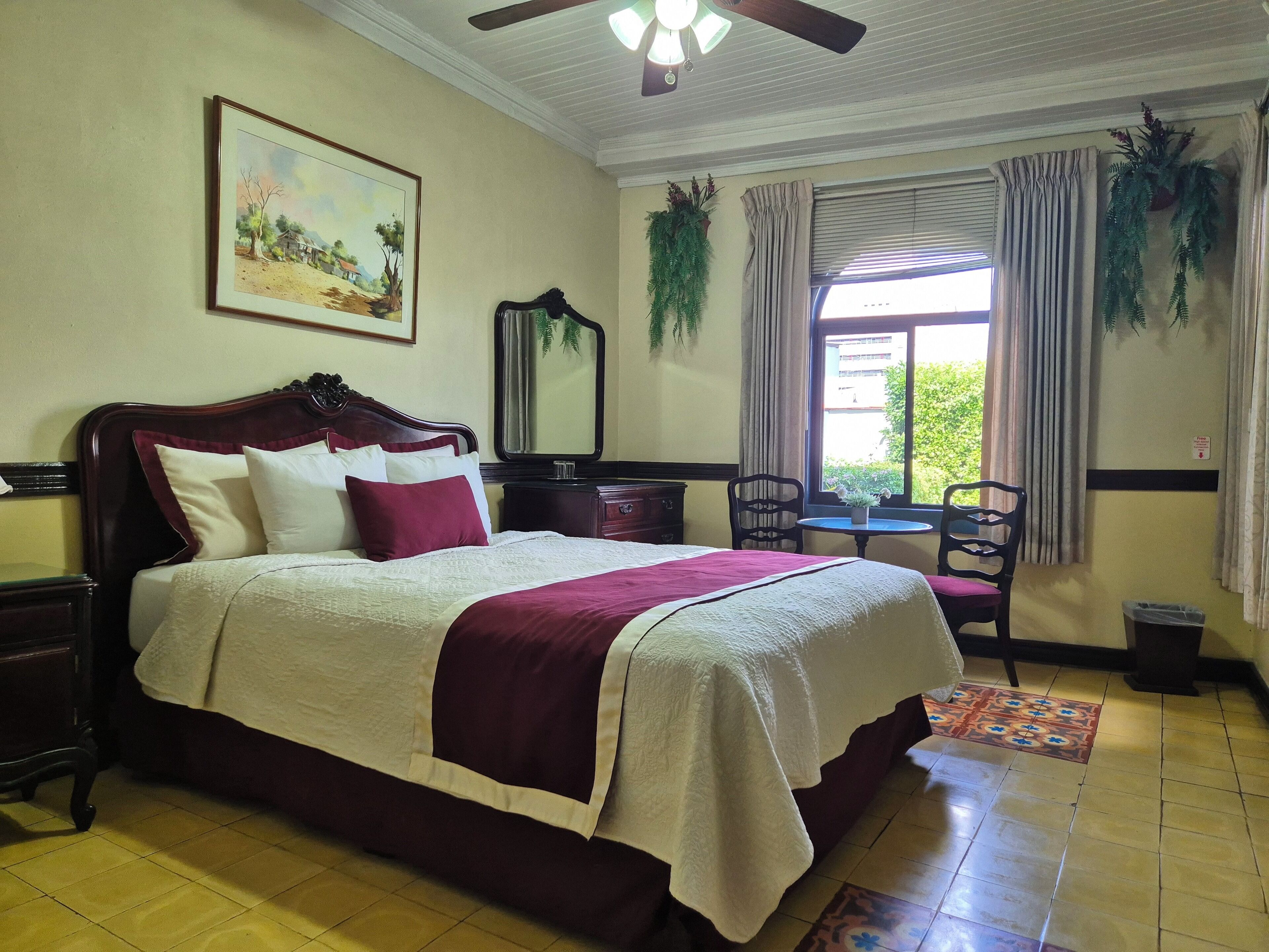 A neatly arranged bedroom with a large bed, a bedside table, a chair, a small table, a ceiling fan, and a window with curtains.