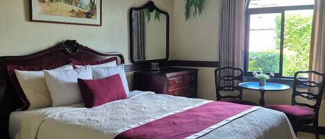 A neatly arranged bedroom with a bed, a ceiling fan, a window with curtains, a small table with chairs, and a painting on the wall.