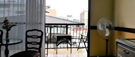 A room with a sliding glass door leading to an outdoor balcony, a standing fan, a small round table with chairs, and a dresser with drawers.