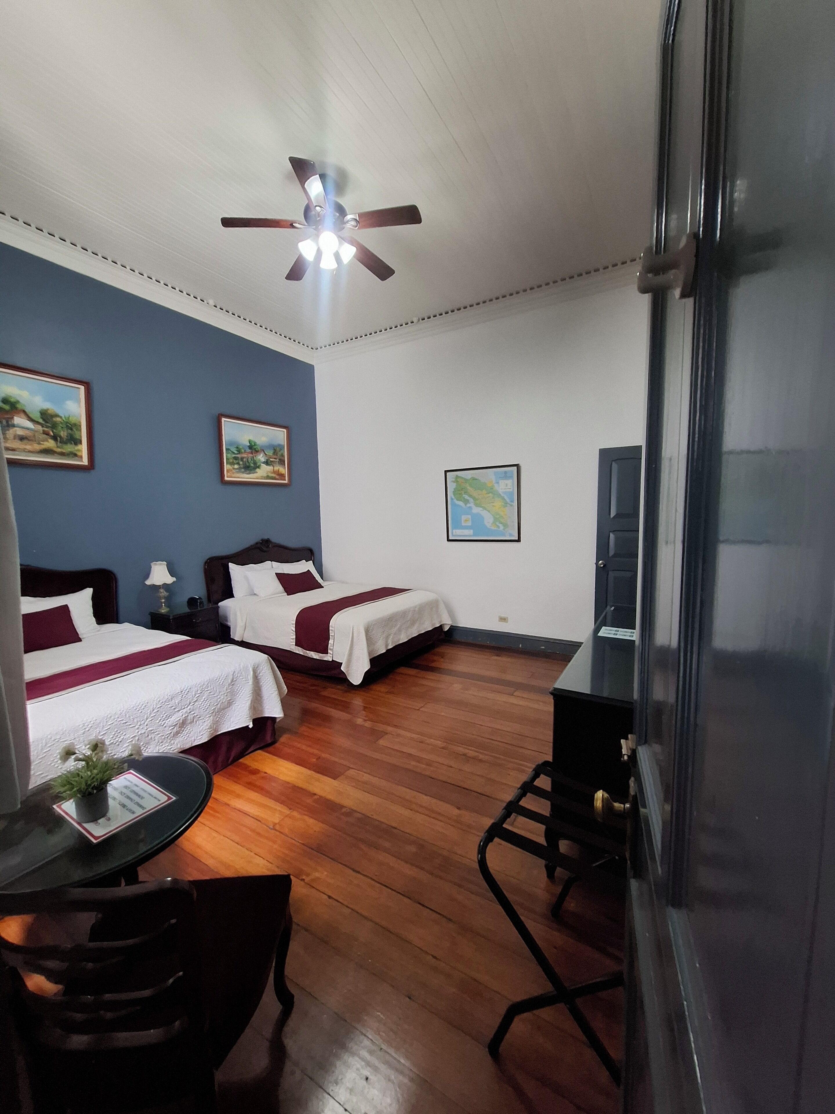 A hotel room with two beds, a desk, a chair, a ceiling fan, and a map on the wall.