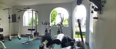 Academia com diversos equipamentos de exercício, janelas amplas e vista para o verde lá fora.