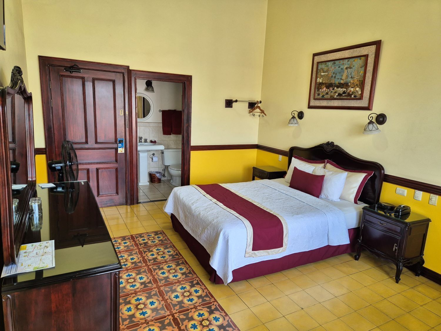 A hotel room with a bed, bedside table, dresser, and a bathroom visible through an open door.