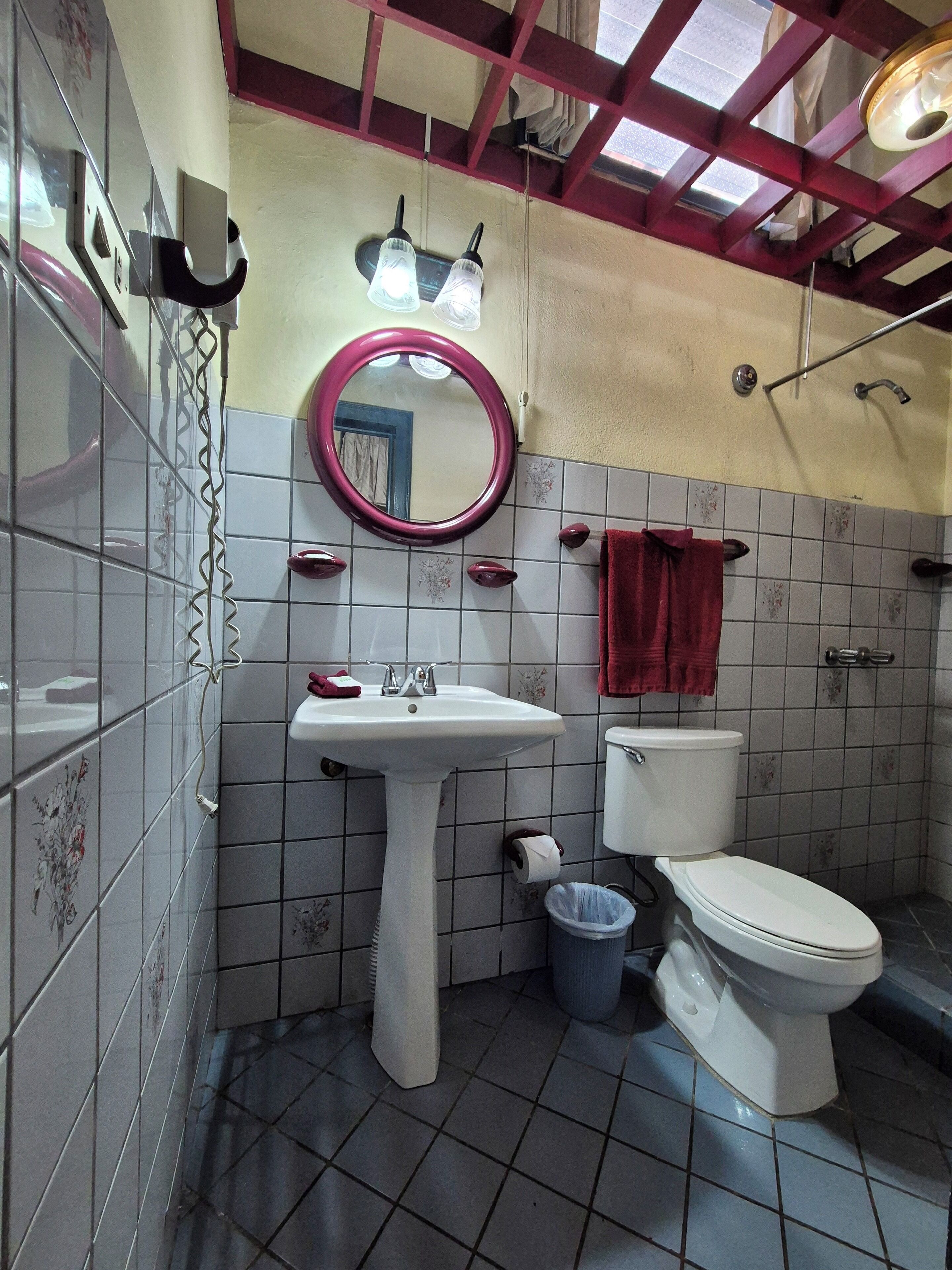 A bathroom with a pedestal sink, a toilet, a trash bin, a red towel, and a mirror.