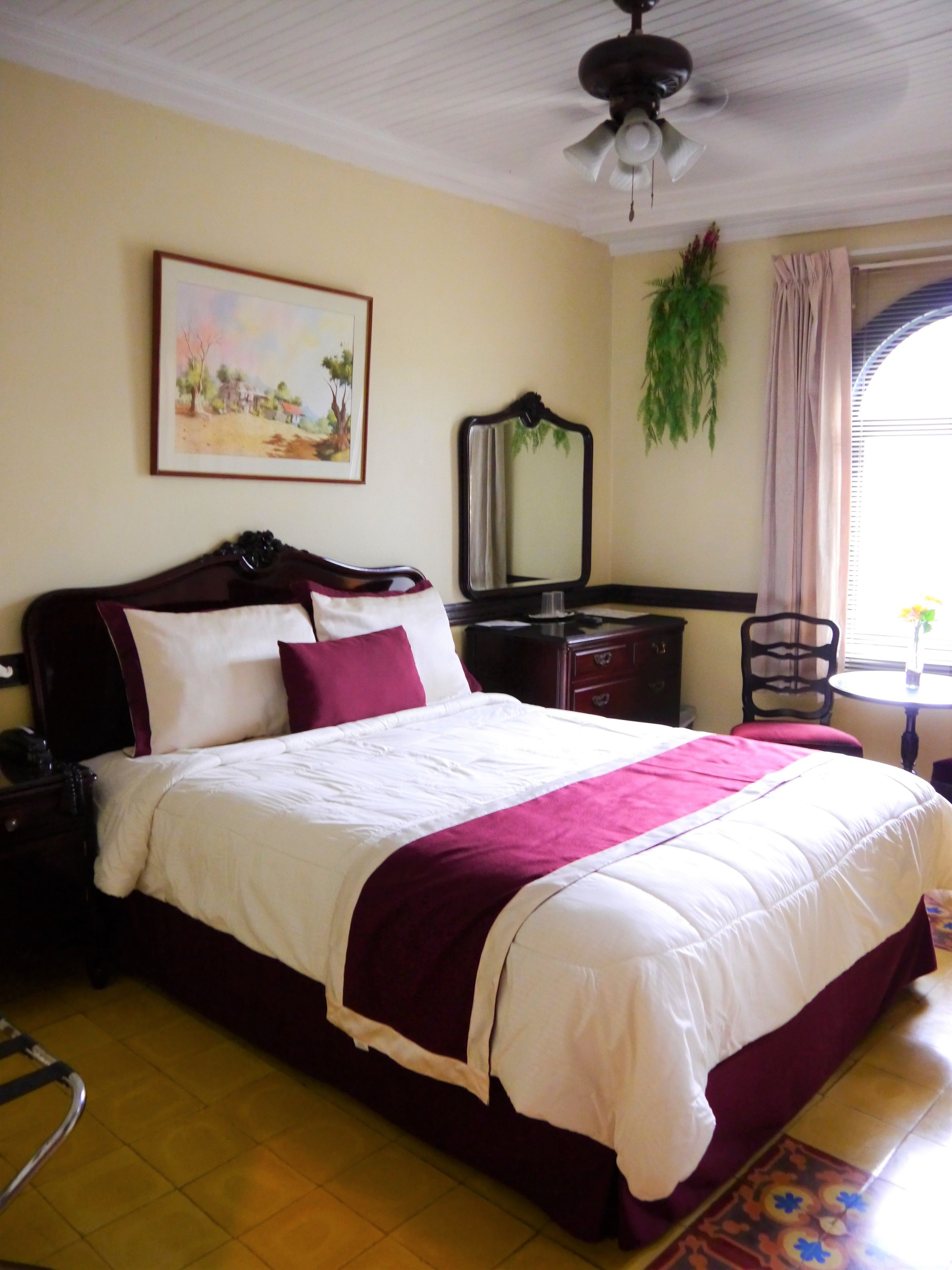 A neatly made bed with a white and maroon bedspread, a wooden headboard, a nightstand with a telephone, a desk with a mirror, and a window with curtains.