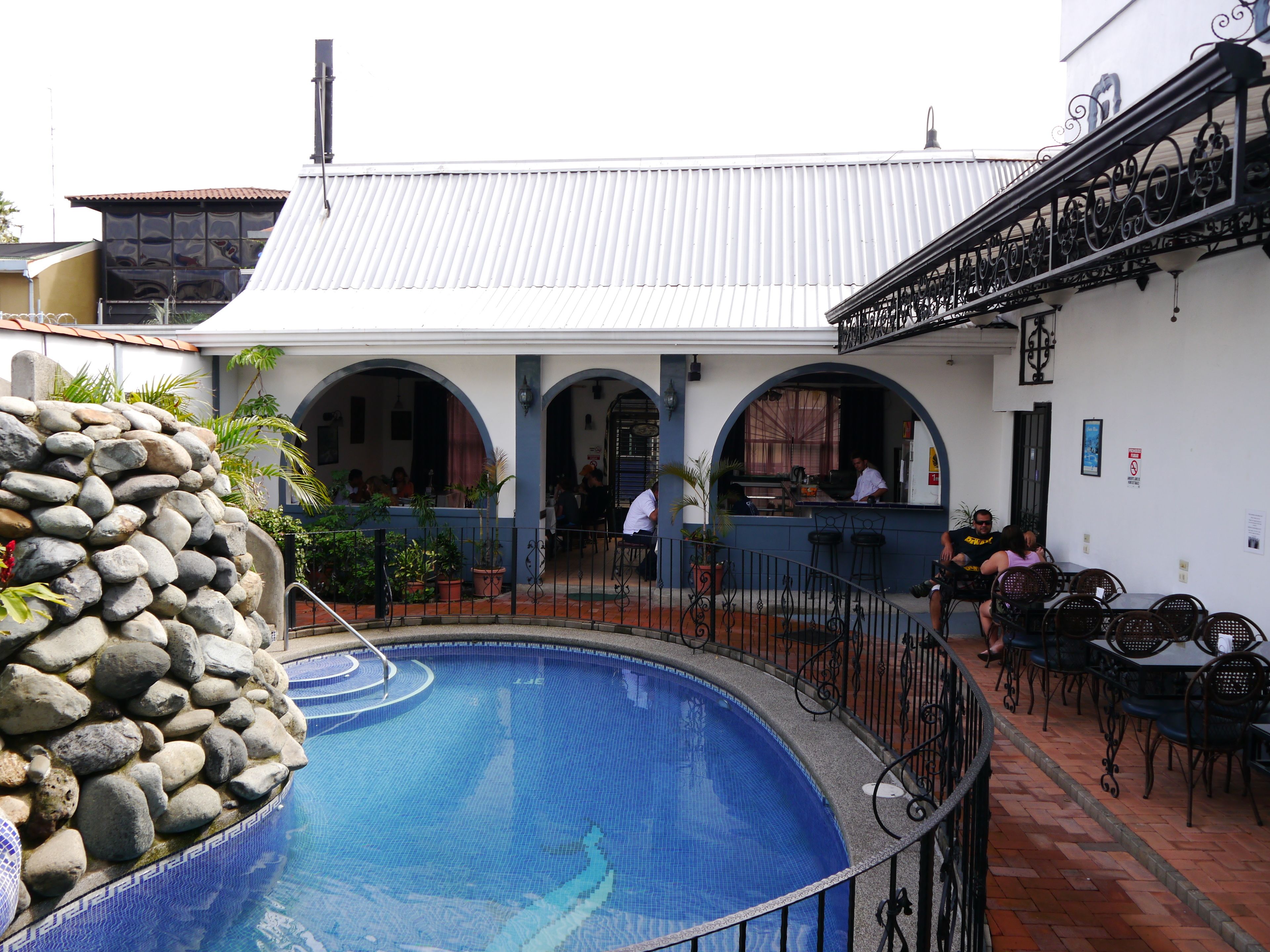A swimming pool with a stone wall, a restaurant with outdoor seating, and a building with a metal roof.