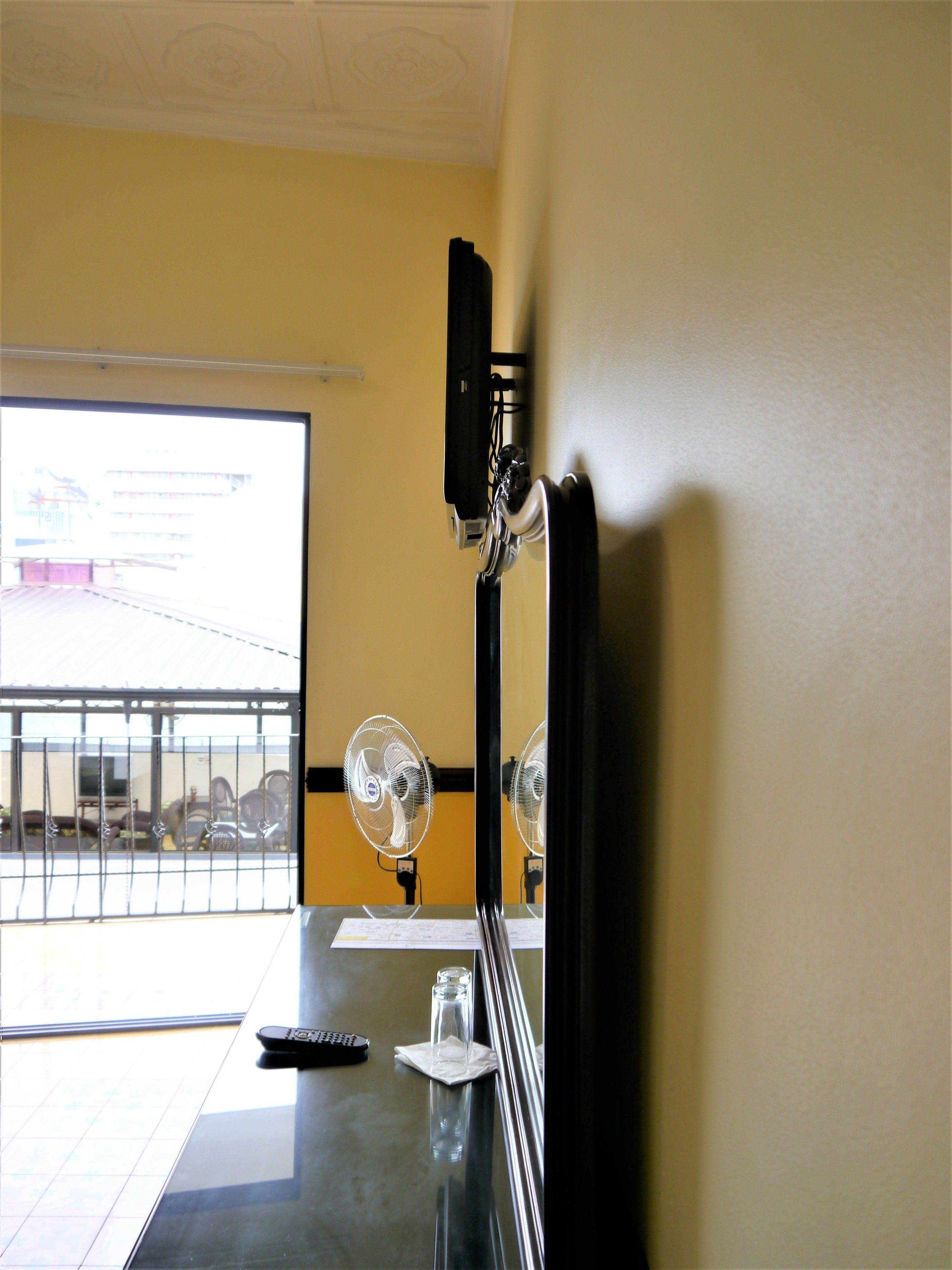 A hotel room with a large window, a desk with a remote control, a glass of water, and a fan.