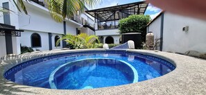 Una piscina con borde de azulejos, rodeada por un solárium de piedra, con un edificio al fondo.