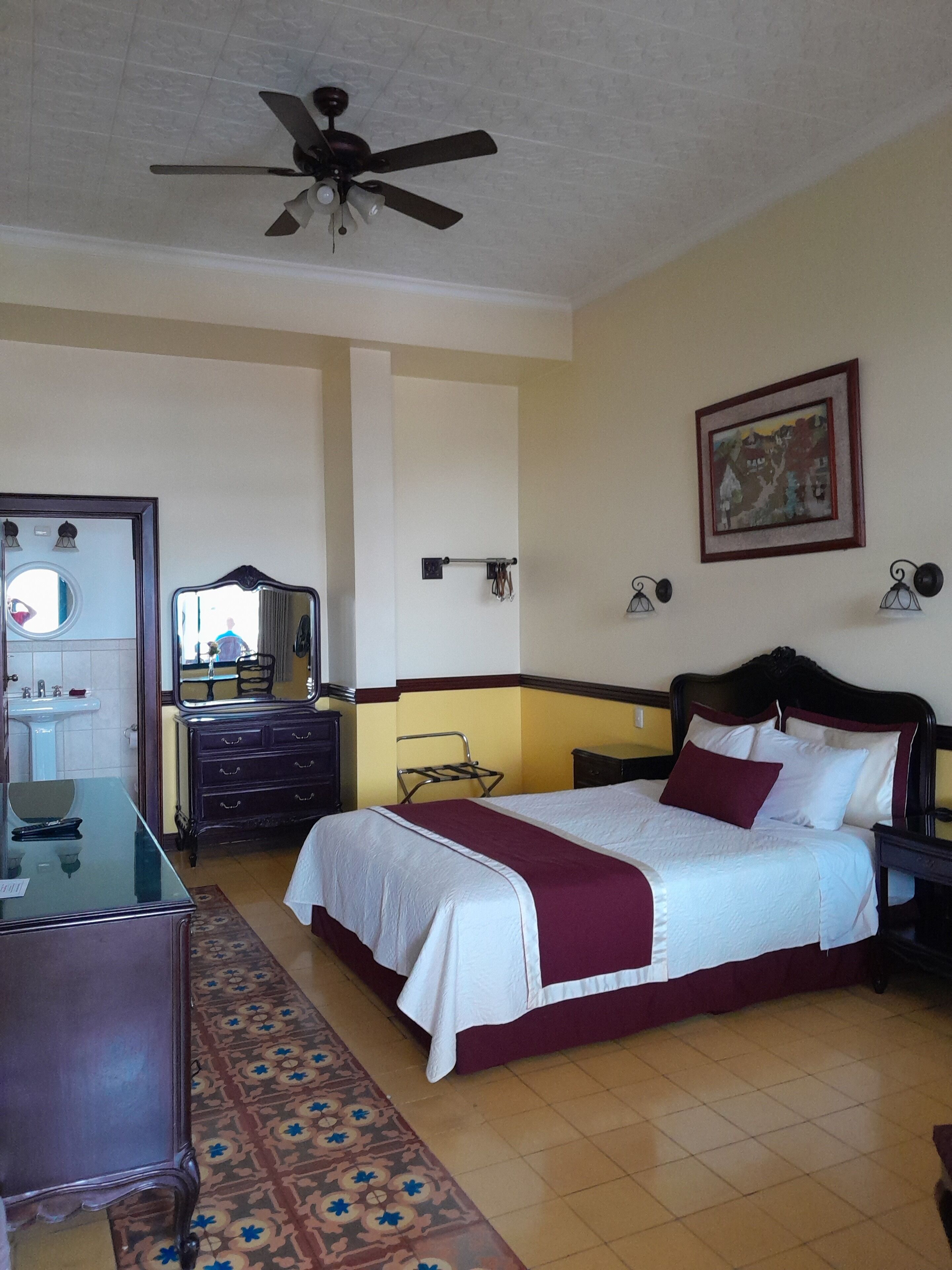 A hotel room with a bed, a dresser, a vanity with a mirror, a ceiling fan, and a painting on the wall.