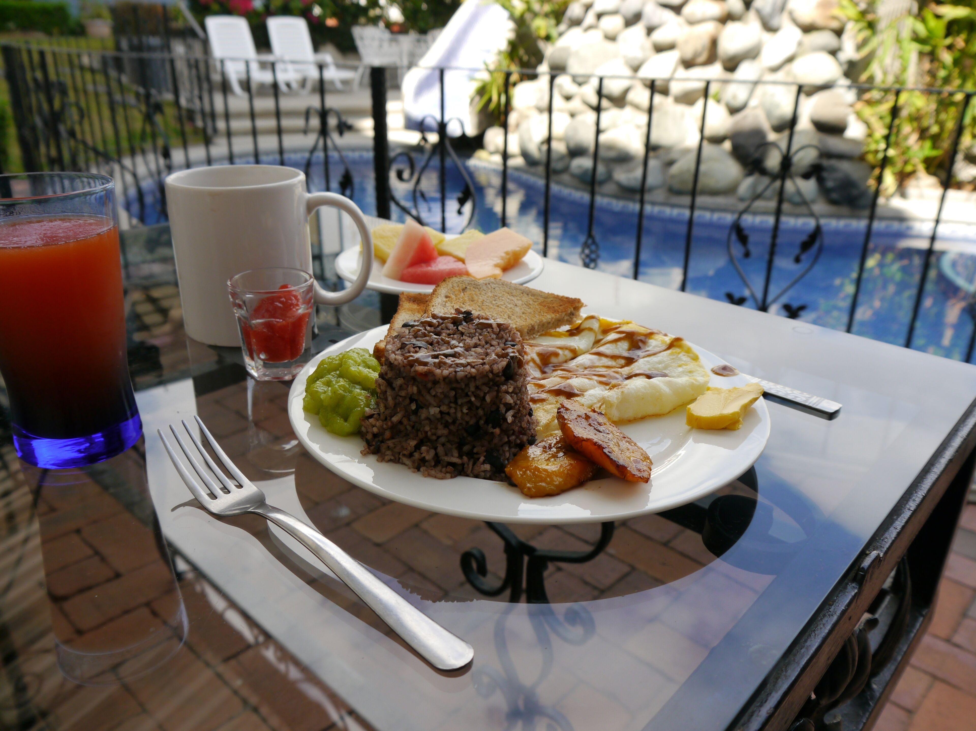 A breakfast plate with toast, eggs, and a brown dessert, a glass of iced tea, and a small cup of jam on a table by a pool.