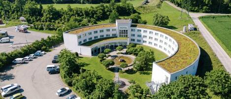 A modern hotel with a curved roof, surrounded by a parking lot and greenery.